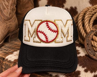 May include: A black and white trucker hat featuring the word MOM in white chenille letters with gold glitter trim. The letter O is replaced by a white baseball graphic with red stitching. The hat has a solid black brim and mesh back panels.