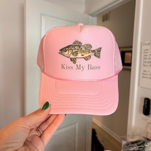 May include: Pink trucker hat with a mesh back and a curved brim. The hat features a graphic of a fish and the text "Kiss My Bass" in white. The hat is a light pink color.