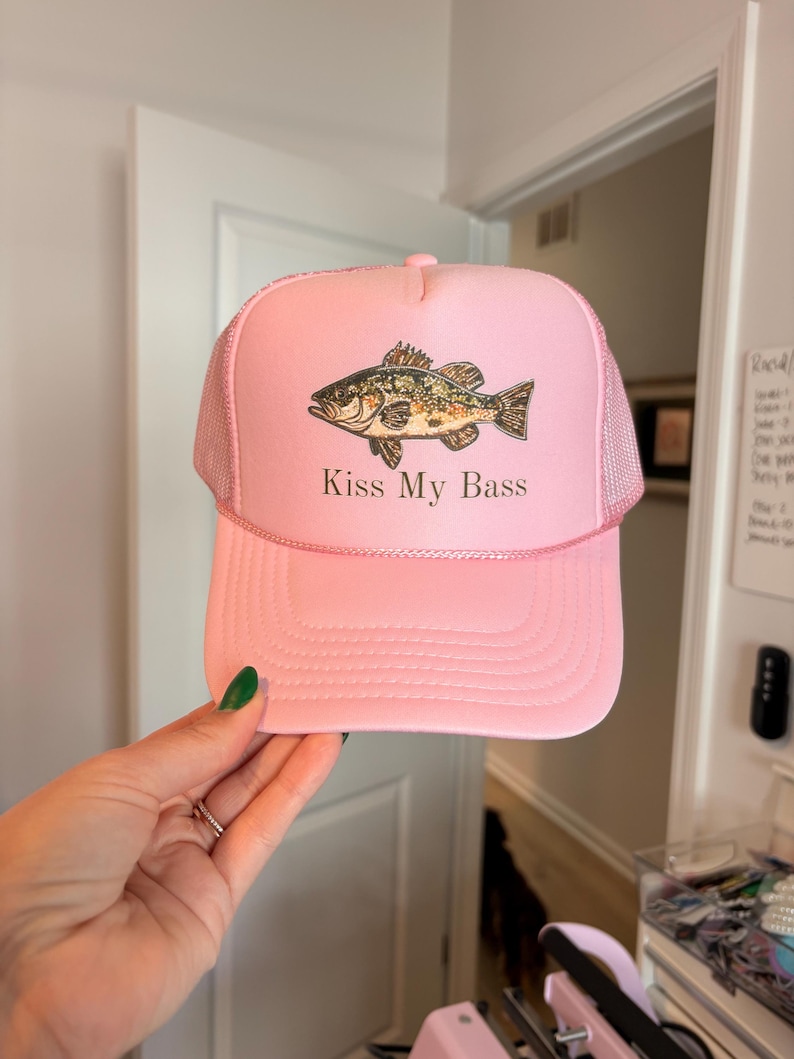 May include: Pink trucker hat with a mesh back and a curved brim. The hat features a graphic of a fish and the text "Kiss My Bass" in white. The hat is a light pink color.
