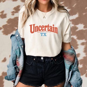 Może przedstawiać: Kremowy t-shirt z krótkim rękawem z czerwonym napisem "Uncertain" nad niebieskim napisem "TX". Koszulka jest noszona z czarnymi szortami dżinsowymi i niebiesko-czerwono-szarą wzorzystą kurtką dżinsową narzuconą na ramiona.