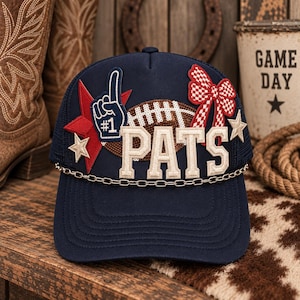 May include: A navy blue trucker hat featuring the word PATS in large beige letters, a sequined football, a red star, a blue foam finger patch, a red and white checkered bow, and a silver chain trim across the base of the front panel.