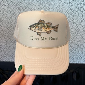 May include: Tan trucker hat with a fish graphic and the text "Kiss My Bass". The hat features a mesh back and a curved brim. The fish is brown, green, and black.