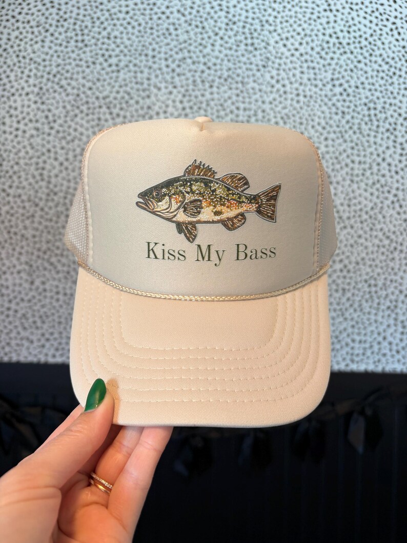 May include: Tan trucker hat with a fish graphic and the text "Kiss My Bass". The hat features a mesh back and a curved brim. The fish is brown, green, and black.