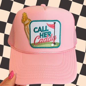 May include: Pink trucker hat with a mesh back and a curved brim. The hat features an embroidered patch with the text "CALL HER CADDY" and a golf-themed design. The hat is held by a hand with pink nail polish.