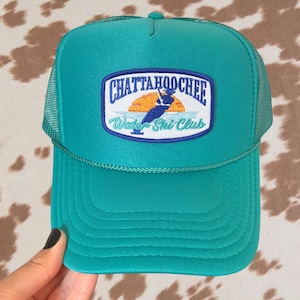 Pode incluir: Um boné trucker verde-azulado com parte traseira em rede e um emblema frontal com o texto CHATTAHOOCHEE Water Ski Club. O emblema apresenta a silhueta azul-escura de um esquiador aquático contra um sol laranja e água azul-clara, com borda azul.