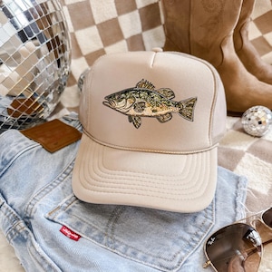 May include: Tan trucker hat featuring a detailed embroidered fish design. The hat has a mesh back and a curved brim. The fish is depicted in shades of green, brown, and gold. The hat is resting on a pair of light-wash denim shorts.