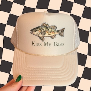 May include: Tan trucker hat with a fish graphic and the text "Kiss My Bass." The hat features a mesh back and a curved brim. The fish illustration is detailed with shades of brown, green, and gold.