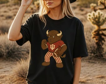 May include: Black t-shirt featuring a cartoon bison in a football uniform. The bison is brown with a red jersey and white and red striped pants. The shirt is worn by a person wearing a black hat and sunglasses.