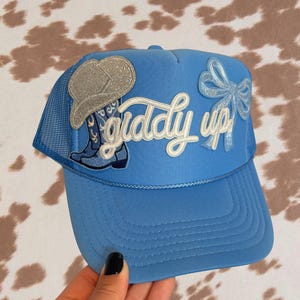Puede incluir: Gorra de camionero azul claro con parte trasera de malla y visera curva. La gorra presenta parches bordados de una bota de vaquero, un sombrero de vaquero y un lazo. Las palabras "giddy up" están bordadas en escritura blanca.