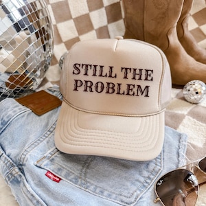 Op de afbeelding: Een beige truckerpet met de tekst "STILL THE PROBLEM" geborduurd in donkerrood. De pet ligt op een lichtblauwe spijkerbroek, met een zonnebril en cowboylaarzen op de achtergrond. Een discobal en een kleine decoratieve bal zijn ook zichtbaar.