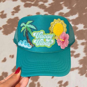 May include: A teal trucker hat featuring embroidered patches of a green palm tree, blue ocean waves, a yellow sun with gold glitter, a pink hibiscus flower, and the text Vacay Vibes in a light blue script font with a bright yellow outline.