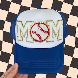 May include: White trucker hat with a blue mesh back. The front of the hat has a gold glitter MOM with a red and white baseball in the center.