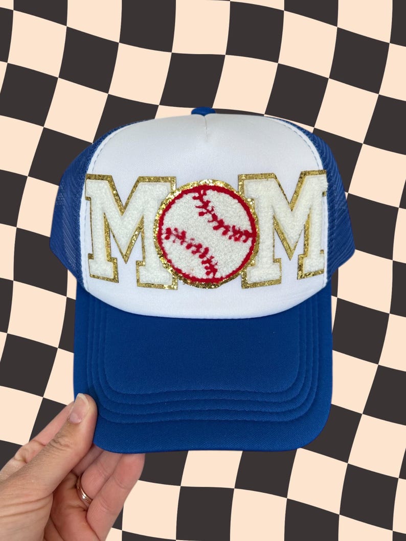 May include: White trucker hat with a blue mesh back. The front of the hat has a gold glitter MOM with a red and white baseball in the center.