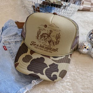 Peut inclure: Casquette de camionneur beige et camouflage avec un motif marron d'un cow-boy à cheval, des cactus et le texte "For a late night call, for a real good time". La casquette a un dos en filet et une visière incurvée.