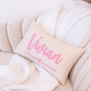 May include: A rectangular beige pillow with the name "Vivian" embroidered in pink, along with a pink heart and line design. The pillow is resting on a beige sofa, with a white blanket in the background.