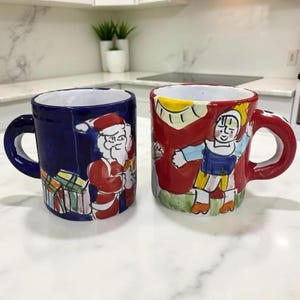 Puede incluir: Dos tazas de cerámica con diseños pintados a mano. Una taza es azul marino con una figura de Papá Noel y regalos envueltos. La otra es roja con una figura y un sol. Ambas tazas tienen un asa curva.