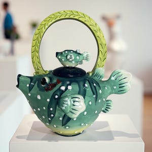 May include: A whimsical, hand-painted ceramic teapot in the shape of a pufferfish. The teapot is primarily green with white polka dots and a yellow base. A small fish figurine sits atop the lid, and a green handle arches over the top. The teapot is displayed on a white surface.