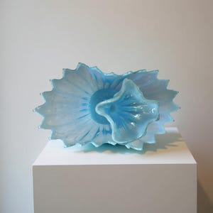 Puede incluir: Escultura de vidrio azul claro con forma de flor, con una forma central en forma de copa rodeada por una capa exterior m&aacute;s grande y con volantes. La escultura se exhibe sobre un pedestal blanco sobre un fondo blanco liso.