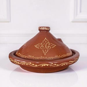 May include: A brown ceramic tagine with a conical lid and a wide circular base. The exterior features hand-painted cream-colored decorative patterns, including a central symmetrical motif on the lid and a wavy line design around the base rim.