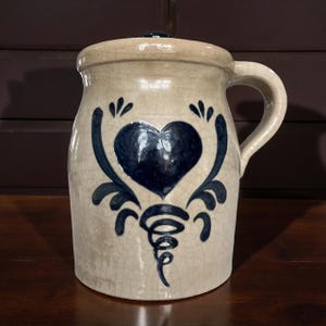 Puede incluir: Una jarra de cerámica de color crema con un corazón azul y un diseño floral. La jarra tiene una tapa y un asa.