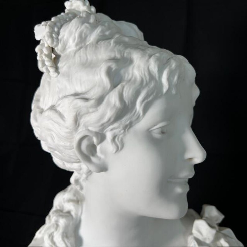 May include: A white porcelain bust of a woman with a detailed hairstyle and a delicate facial expression. The bust is a classic example of fine craftsmanship.