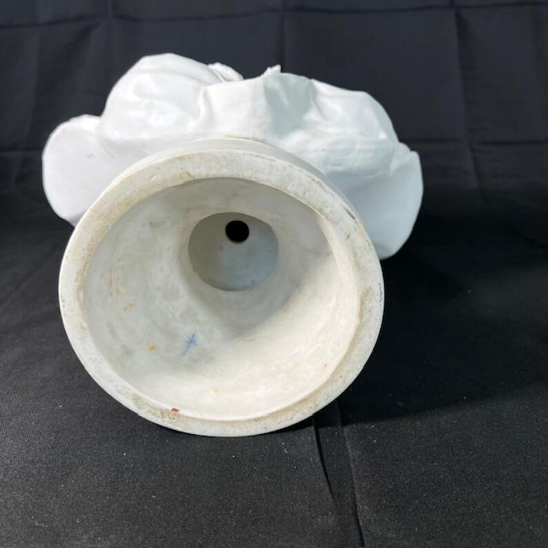 May include: White porcelain bust sculpture with a detailed design. The bust is on a round base with a hole in the center.