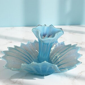 Puede incluir: Centro de mesa de cristal azul claro con dise&ntilde;o floral. El centro de mesa tiene un jarr&oacute;n central y una base ancha en forma de p&eacute;talo. El cristal es transl&uacute;cido, con sutiles variaciones de color y textura.