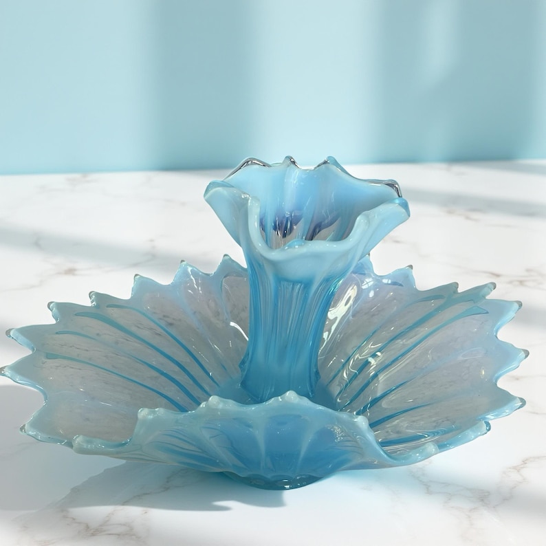 Puede incluir: Centro de mesa de cristal azul claro con dise&ntilde;o floral. El centro de mesa tiene un jarr&oacute;n central y una base ancha en forma de p&eacute;talo. El cristal es transl&uacute;cido, con sutiles variaciones de color y textura.