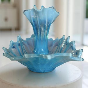 Puede incluir: Centro de mesa de cristal azul claro con un borde ondulado y una base central acampanada. El cristal es transl&uacute;cido. La pieza est&aacute; sobre una superficie blanca.
