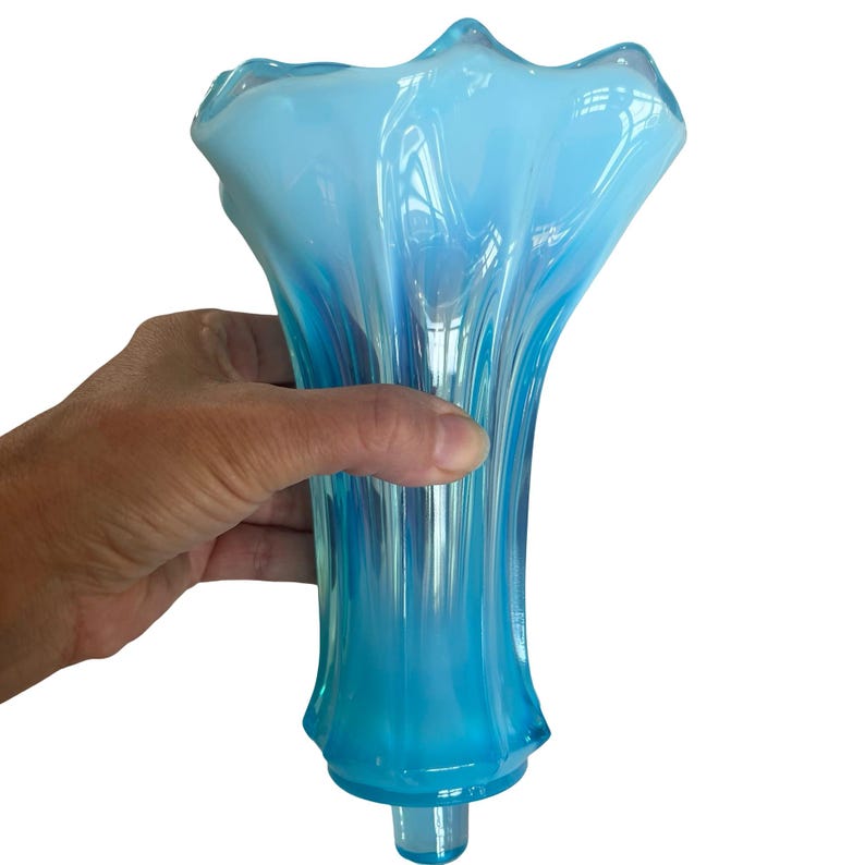 Puede incluir: Un jarr&oacute;n de cristal azul claro con la parte superior con volantes y un cuerpo acampanado y acanalado. El jarr&oacute;n tiene una base cil&iacute;ndrica transparente. El cristal tiene una calidad transl&uacute;cida, permitiendo el paso de la luz. El jarr&oacute;n es sostenido por una mano.