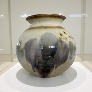 May include: A ceramic vase with a rounded body and a narrow neck. The vase has a cream-colored base with blue, purple, and brown abstract designs. The rim is a darker brown. The vase is displayed on a white surface.