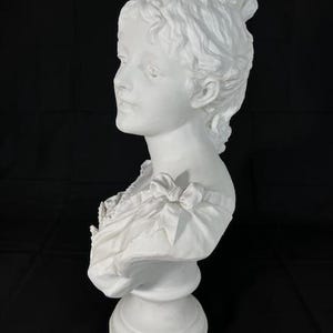 May include: A white porcelain bust of a woman with her hair styled in an updo. The bust is on a pedestal and is in a profile view.