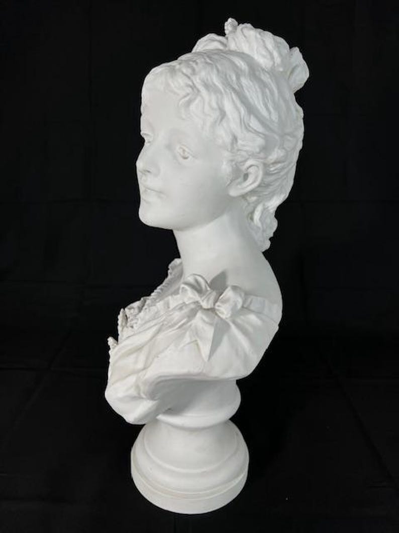 May include: A white porcelain bust of a woman with her hair styled in an updo. The bust is on a pedestal and is in a profile view.