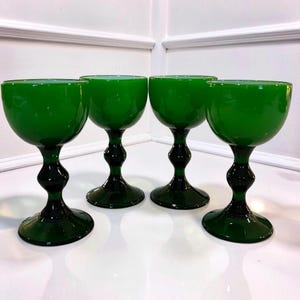 May include: Four green wine glasses with a rounded bowl and a dark green, decorative stem. The glasses are made of glass and are arranged in a row on a white surface. The stem has a bulbous design.