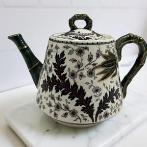 May include: A decorative teapot with a white background, featuring a floral pattern with burgundy and gray accents. The handle and spout are dark green with gold trim. The lid has a gold handle.