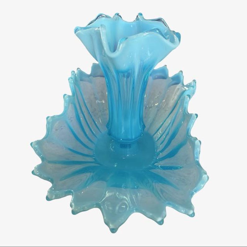 Puede incluir: Centro de mesa de cristal azul claro con borde ondulado. El centro de mesa presenta una estructura central en forma de jarr&oacute;n que se eleva desde una base ancha en forma de p&eacute;talo. El cristal tiene una calidad transl&uacute;cida.