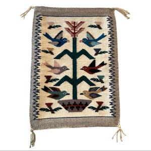 May include: Rectangular woven wall hanging with a cream-colored background and a gray border. The design features a central tree with colorful birds and floral accents. Tassels are attached at each corner. The textile is likely made of wool.