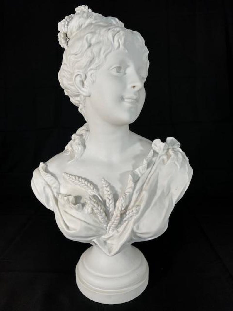 May include: White porcelain bust of a woman with a crown of wheat and a draped cloth. The bust is on a white pedestal.