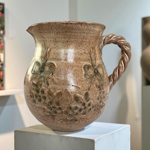 Puede incluir: Jarra de cerámica moteada de color beige con asa trenzada. La jarra presenta diseños florales y lazos en relieve en tonos verdes y marrones. La jarra se exhibe sobre un pedestal blanco.