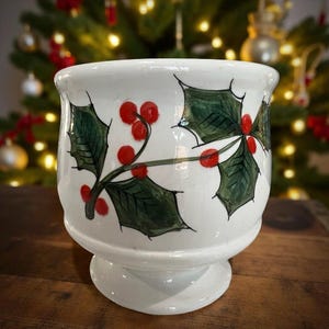 May include: White ceramic footed bowl with a hand-painted holly design. The bowl features green holly leaves and red berries, perfect for holiday decor. The bowl is on a wooden surface.