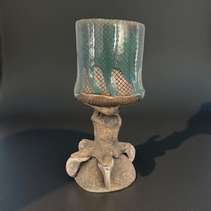 Puede incluir: Una taza para beber de cerámica con un esmalte verde y un diseño texturizado. La taza está unida a una base esculpida que se asemeja a una pata de pájaro.
