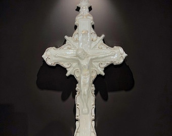 May include: A white ceramic crucifix featuring a glossy finish with gold-toned trim along the edges. The cross displays an ornate, scrolled design with a star at the top and a central figure of Jesus with arms outstretched in a traditional pose.