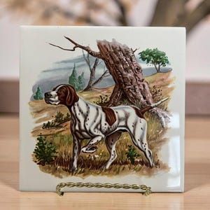 May include: Square ceramic tile featuring a Pointer dog illustration. The dog is white with brown spots and a brown head, standing in a grassy field with a tree and a cloudy sky in the background. The tile is displayed on a gold-colored metal stand.