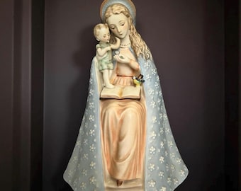 May include: A ceramic statue of a woman with a child, wearing a light blue cloak with white floral patterns. The woman holds a small bird and a small object. The statue is on a wooden base and has a halo.