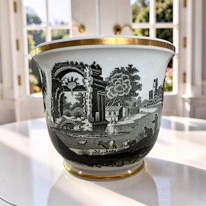 May include: A white ceramic planter with a black and white pastoral scene. The planter has a gold rim at the top and base. The design features buildings, trees, and figures. The planter sits on a white surface.