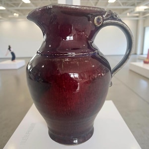 May include: A burgundy ceramic pitcher with a silver handle and spout. The pitcher has a glossy finish and a bulbous body. The handle is adorned with decorative elements. The pitcher is displayed on a white surface.