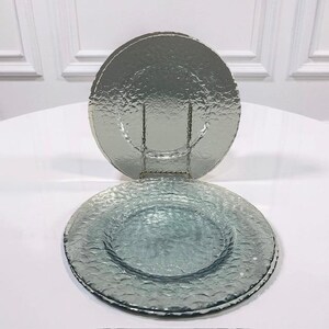 May include: Two clear, textured glass charger plates in a light blue-green hue. One plate is resting on a metal stand, while the other is lying flat. The plates have a textured surface and are suitable for decorative use.