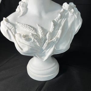 May include: White ceramic bust sculpture of a woman with a draped fabric and wheat stalks. The bust is on a pedestal base.