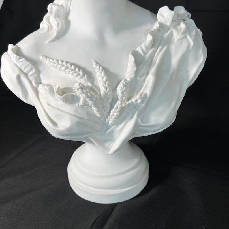 May include: White ceramic bust sculpture of a woman with a draped fabric and wheat stalks. The bust is on a pedestal base.