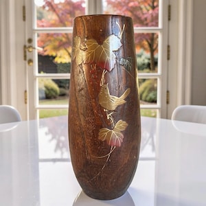 May include: A tall, wooden vase with a natural wood grain finish. The vase is adorned with gold-colored leaf and bird designs. The leaves have red accents. The vase is cylindrical with a slightly flared top, suitable for floral arrangements or decorative display.